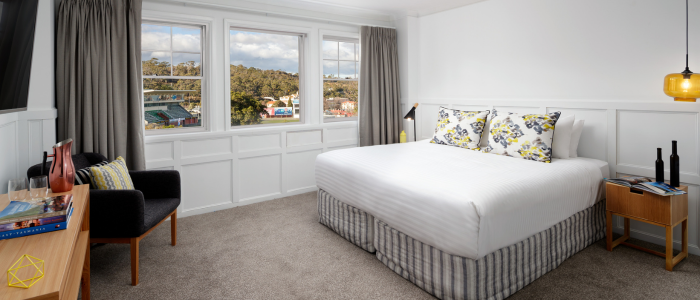 Well-lit bedroom with a large bed featuring white linens and floral pillows, positioned against the right wall. A wooden nightstand with bottles and books sits beside the bed under a hanging yellow lamp. An armchair with a striped cushion and a small table with books and decor are on the left. The room has three large windows with gray curtains, white paneled walls, and light brown carpet.