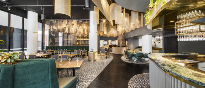 A modern, stylish restaurant interior featuring black ceilings and floors contrasted by white columns and patterned tiles. The space includes green upholstered booths and chairs, wooden tables, and a curved bar area with hanging glassware. Large cylindrical light fixtures hang from the ceiling, and greenery is incorporated above the bar, creating an elegant and contemporary ambiance.