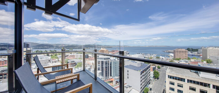 A high-rise balcony with two modern lounge chairs facing outward, overlooking Hobart and a distant body of water. The balcony features glass railings for an unobstructed view of the buildings below and the scenic horizon. The sky is partly cloudy with patches of blue.