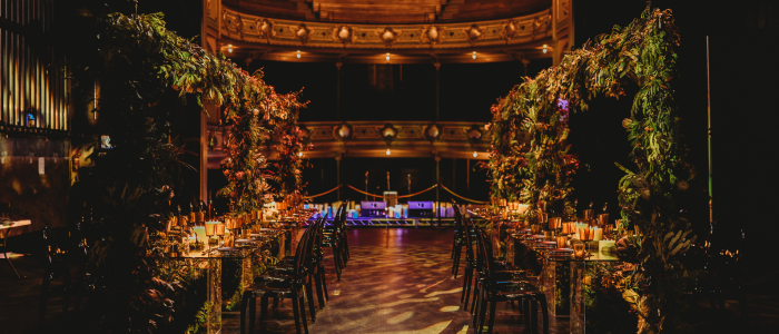 A beautifully decorated banquet hall with two long tables adorned with candles and floral arrangements. The tables are set under arches covered in greenery and flowers, creating a lush, garden-like atmosphere. The background features an ornate balcony with warm lighting, adding to the elegant ambiance of the venue.