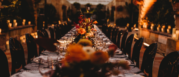 A long, elegantly set dining table in an outdoor or semi-outdoor setting, adorned with floral centerpieces and surrounded by black chairs. Warm ambient lighting and candles along the sides create a cozy and inviting atmosphere.