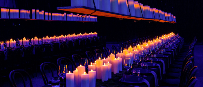 A long dining table set for an event, featuring numerous lit candles along its center and suspended platforms above holding additional candles. The dim lighting creates a warm, intimate atmosphere, with neatly arranged chairs, wine glasses, and place settings visible.