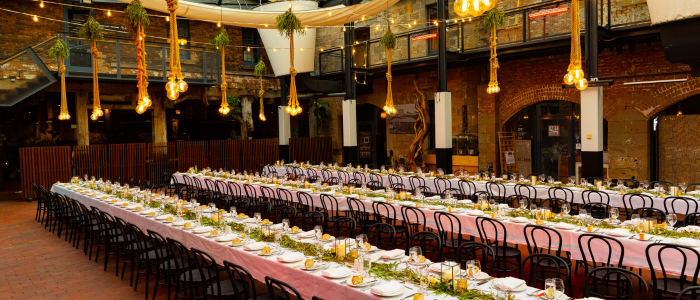 A large, elegantly decorated dining area set for an event, featuring three long tables with white tablecloths, greenery, and candles. Black chairs surround the tables, while hanging light bulbs and suspended plants enhance the ambiance. The background includes exposed brick walls and a mezzanine level with extra seating or standing space.