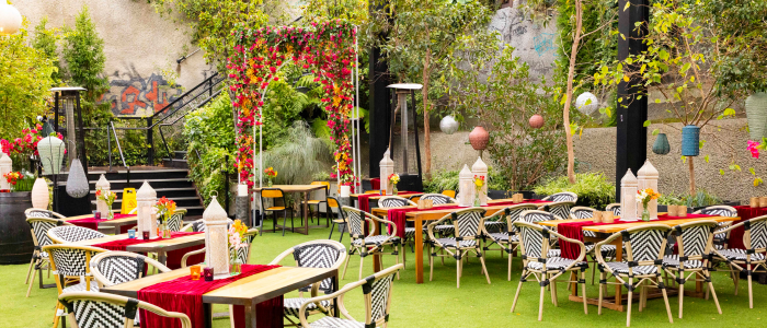 An outdoor dining area with tables covered in red tablecloths and decorated with white lanterns, set on green artificial grass. A floral archway with red and yellow flowers stands in the background, surrounded by lush plants and trees. Hanging lanterns and lights enhance the ambiance, with a staircase partially hidden by greenery leading to another level.