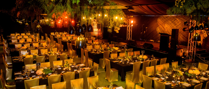 Elegantly decorated event space with rows of rectangular tables set for a formal dinner, featuring floral centerpieces, gold-covered chairs, and warm lighting. A stage with microphones and speakers is visible in the background.