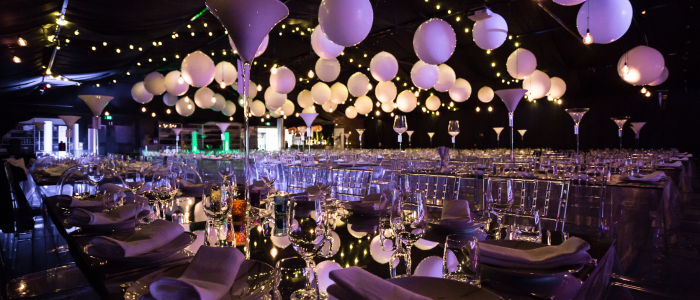 Sophisticated event space decorated with round white balloons and string lights hanging from the ceiling. Tables are set with glassware, plates, napkins, and centerpieces, creating a festive ambiance.