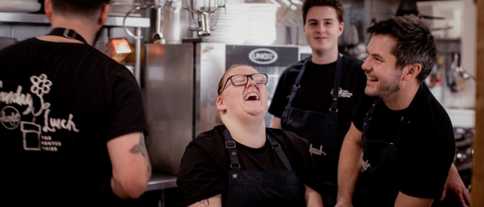 Four people in a kitchen setting, likely restaurant staff, wearing black shirts and aprons. Center person laughing and others smiling. The background includes kitchen equipment such as an oven and various utensils.