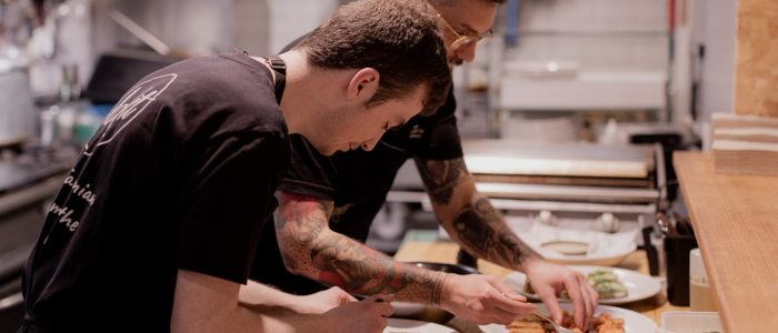Two individuals working together in a kitchen, preparing or plating food on a counter. The background includes kitchen equipment and utensils. One person wears a black shirt with text on the back, and the other wears glasses.