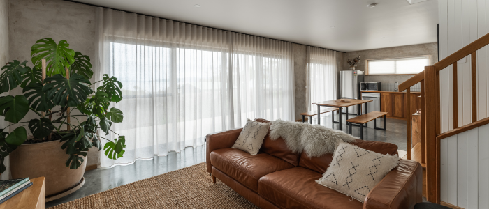 Modern living room featuring a large brown leather sofa with white and patterned cushions and a fur throw, a potted plant to the left, sheer floor-to-ceiling curtains, and an open kitchen with wooden cabinets and a dining table in the background. A staircase with wooden steps and white paneling is visible on the right, with a woven rug on the polished concrete floor.