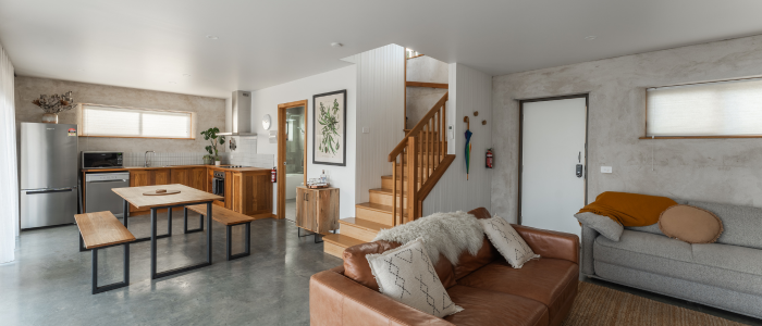 Modern open-plan living area with a kitchen featuring wooden cabinets and stainless steel appliances, a central wooden dining table with benches, and a living room with a brown leather sofa and a gray sofa, both adorned with cushions and throws. A staircase leads to the upper floor, and the walls are decorated with framed artwork and hanging hooks.