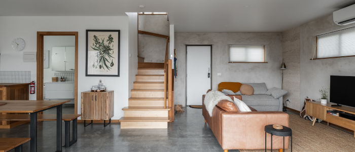 Modern open-plan living room with a central wooden staircase, a dining area to the left featuring a wooden table and benches, and a living area to the right with a gray sofa, brown leather armchair, and a flat-screen TV on a wooden stand. The walls are white and light gray, decorated with botanical artwork, and an air conditioning unit is mounted near the ceiling.
