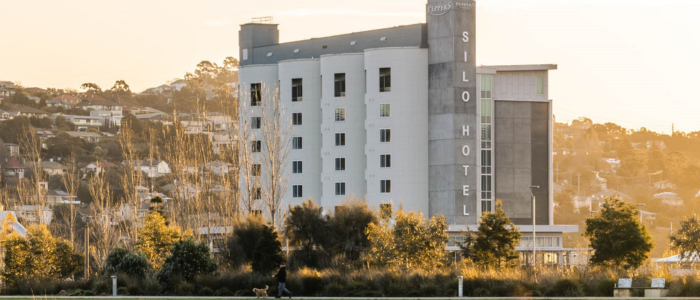 Peppers Silo Hotel in Launceston, a multi-story building with modern architecture, large windows, and a cylindrical tower displaying the words 'SILO HOTEL' vertically. The hotel is surrounded by trees and greenery, with a person walking a dog in the foreground and residential houses on a hillside in the background under an overcast sky.