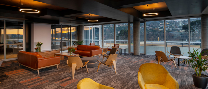 Interior lounge area of the Peppers Silo Hotel in Launceston, featuring large windows with views of a hilly landscape. The room includes orange and yellow chairs, orange sofas, small tables, potted plants, and modern ceiling lighting with recessed and circular fixtures.