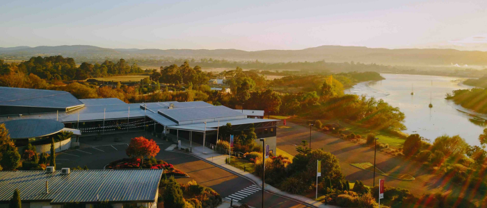Aerial view of a scenic landscape with a modern building complex surrounded by greenery. The Tamar River runs along the right side, with hills and mountains in the background. The low sun casts long shadows and a warm, golden light across the scene.