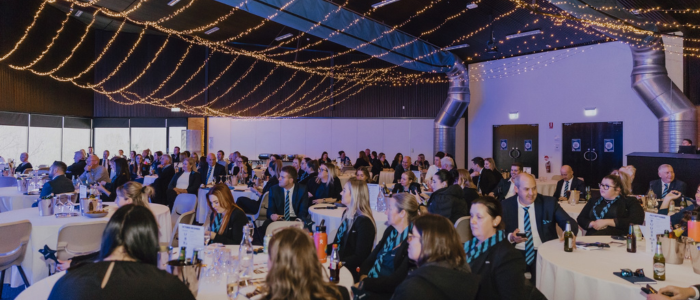 Large indoor event with many people seated at round tables, likely a formal gathering or conference. String lights hang from the ceiling, adding a festive atmosphere.