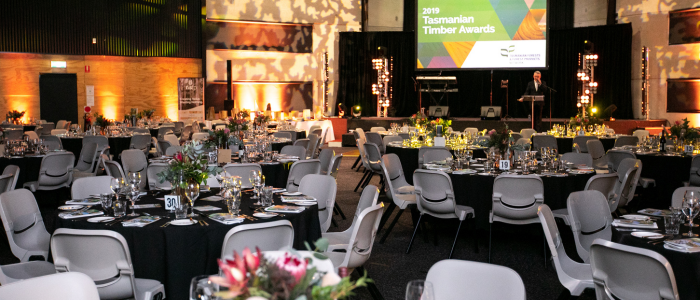 Formal event setup with round tables covered in black tablecloths, set with plates, glasses, and floral centerpieces. A stage in the background displays '2019 Tasmanian Timber Awards' with a person at a podium. The room features warm lighting and an elegant ambiance.