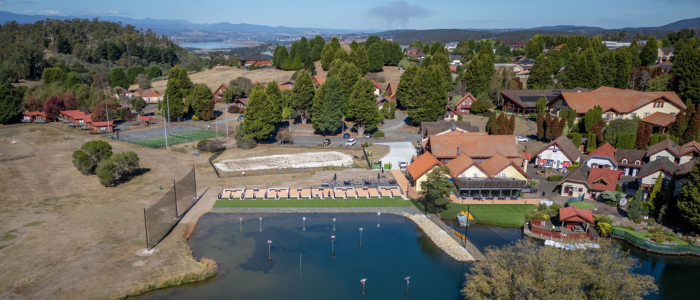 Aerial view of Tamar Valley Resort with red and orange-roofed buildings surrounded by trees. Hills and distant mountains visible under a clear sky.