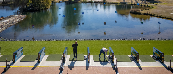 Golf driving range with multiple hitting bays, where two people are practicing swings. One is preparing to hit while the other adjusts the ball or tee. The range features a water hazard with distance markers and is surrounded by greenery and trees.