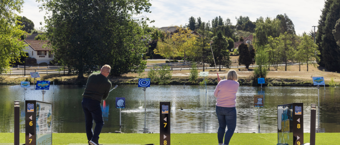 Two people standing on a grassy area near a body of water, aiming at floating targets with various designs and numbers. Trees, houses, and a clear sky form the background.