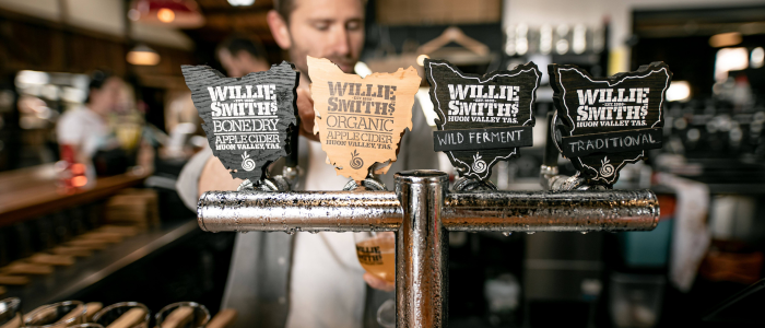 Four taps for Willie Smith's cider at a bar, labeled "Bone Dry Apple Cider," "Organic Apple Cider," "Wild Ferment," and "Traditional," with a blurred background showing the bar interior and patrons.
