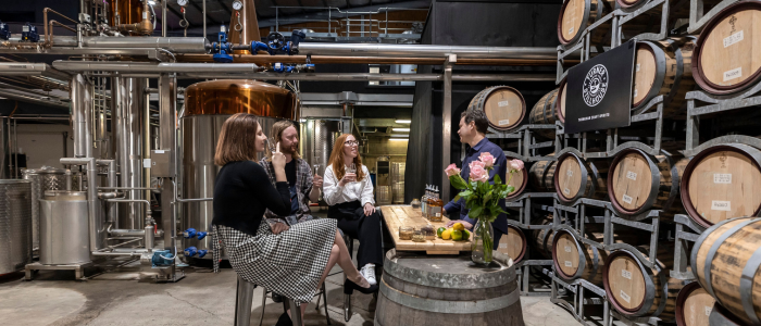our people seated around a table in a distillery or brewery setting, with large metal tanks and barrels in the background. The table is decorated with flowers, fruits, and bottles. A 'Brewery Tours' sign is visible on the right.