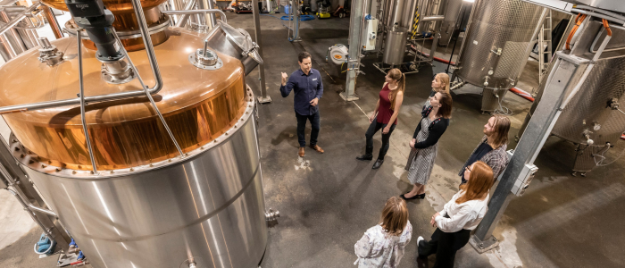 Group of people standing in a brewery or distillery, gathered around large stainless steel and copper tanks. One person appears to be leading a tour or educational session, with industrial equipment like pipes and valves visible in the background.