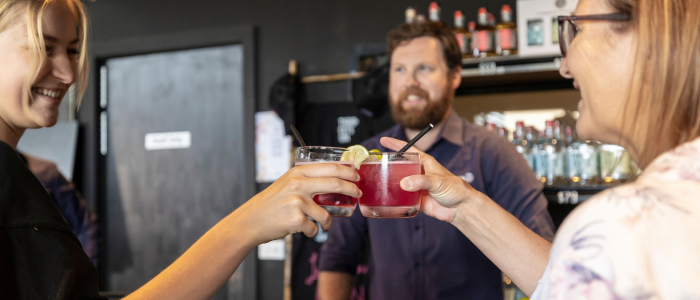 Three people toasting with glasses of red drinks in front of a bar area with bottles on shelves and a closed door in the background.