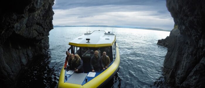 A boat with a yellow inflatable hull and white roof carrying several people is positioned inside a cave or tunnel, surrounded by rocky walls. The view extends out to an open body of water under a cloudy sky.