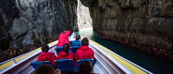 A group of people wearing red jackets sit in a boat navigating through a narrow passage between tall, rocky cliffs. The water is visible below, and the boat appears to be heading toward an opening where light is coming through.