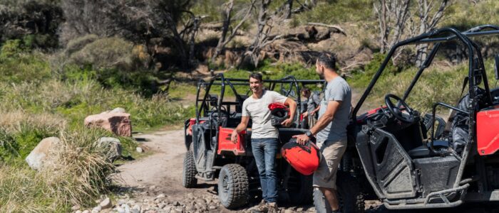 Two people standing next to off-road vehicles on a rocky path in a natural setting. One person is holding a red fuel container, and they appear to be engaged in conversation. The background includes trees and bushes, suggesting an outdoor environment during an off-road adventure.