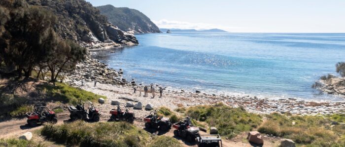 A scenic coastal area with clear blue water and a rocky shoreline. Several off-road vehicles are parked on a dirt path near the beach, and a group of people stand near the water's edge. Tree-covered hills and mountains extend into the background.