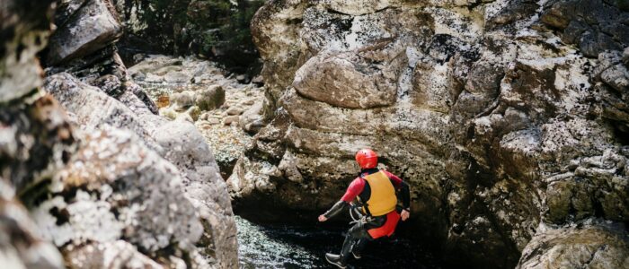 A person wearing a red helmet, yellow life jacket, and black wetsuit is jumping into a narrow rocky gorge with water at the bottom. The area is surrounded by rugged rock formations and greenery in the background.