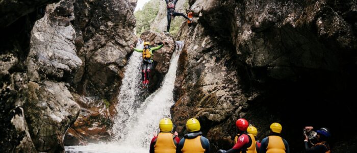 A group of seven people wearing helmets and life jackets are standing in the water at the bottom of a small waterfall, likely participating in canyoning or cliff jumping. Two individuals are mid-air, jumping from the top of the waterfall into the pool below, surrounded by rocky cliffs and flowing water.