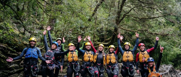 A group of people standing in a shallow body of water, surrounded by lush green trees. They are wearing wetsuits, helmets, and life jackets, likely participating in an outdoor adventure activity such as canyoning or river trekking. The individuals have their arms raised and appear to be posing for the photo.