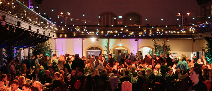 A large indoor event with guests seated at tables and some standing, set in a spacious venue with high ceilings and a visible upper balcony. The room is decorated with string lights, plants, and greenery, creating a warm and festive atmosphere.