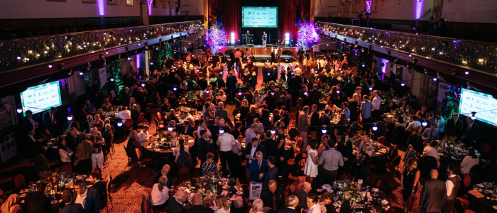 A formal gala event in an ornate hall filled with round tables and guests in formal attire. The room features decorative lighting, a stage with two large screens, and purple-lit trees flanking the stage. Attendees are seated and mingling in a festive, elegant atmosphere.
