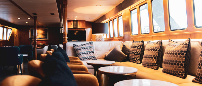 Interior of a luxurious yacht featuring a long beige sofa with patterned cushions, round white marble-topped tables, cushioned chairs, large windows allowing natural light, polished wooden walls and ceiling, a side table with a lamp, and artwork in the background.