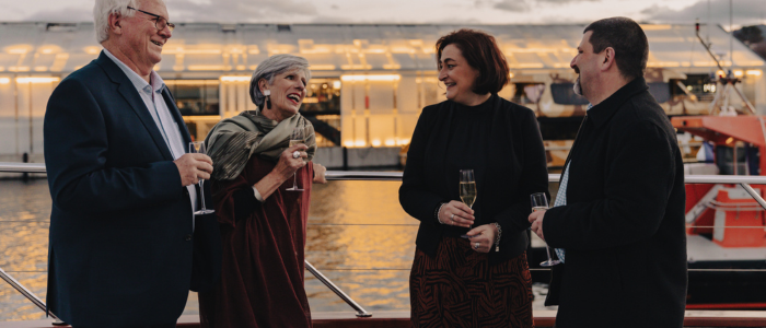 Four people standing on a boat or dock, holding champagne glasses and conversing. In the background, a large illuminated building reflects on the water.