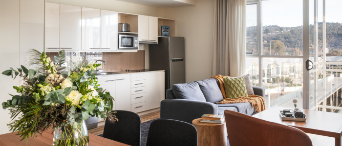 Modern apartment-style living space at Peppers Seaport Hotel featuring an open-plan kitchen with white cabinets, microwave, stove, and refrigerator. The living room includes a grey sofa with cushions near large windows offering views of buildings and greenery. A dining table with chairs and a floral centerpiece sits in the foreground, all illuminated by natural light.