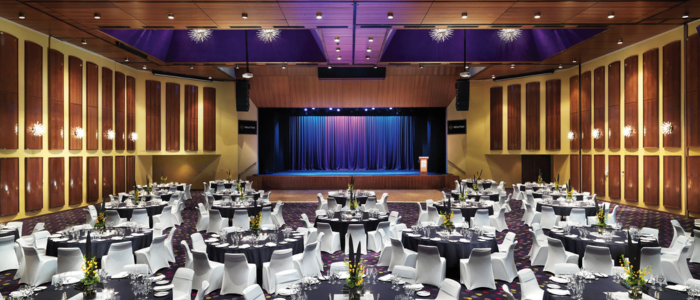 Elegant banquet hall with round tables covered in black tablecloths and surrounded by white chairs. Each table has floral centerpieces and formal place settings. A stage with blue curtains and a podium is visible in the background. The room features wooden wall panels, modern light fixtures, and a decorative ceiling with recessed lighting.