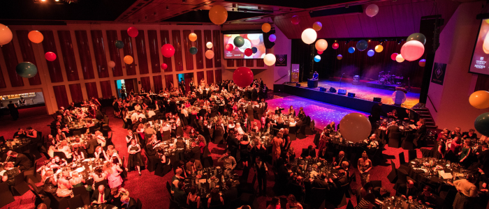 Festive event at Wrest Point Hotel & Conference Centre with round tables filled with guests dining and socializing. Colorful balloons float above the tables, and a stage with lighting and musical equipment is set up in the background. Two large screens flank the stage, displaying visuals related to the event.