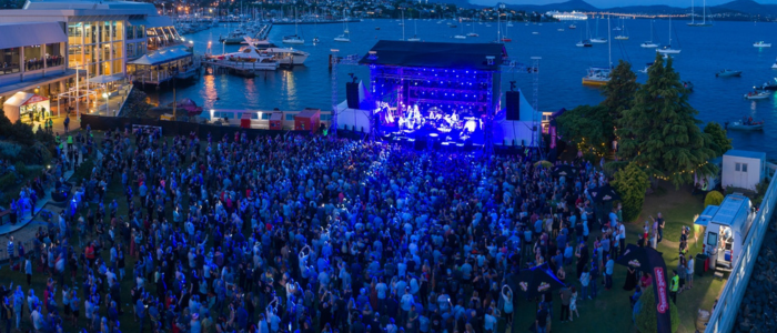 Outdoor concert at Wrest Point Hotel & Conference Centre near the waterfront during twilight. A brightly lit stage with performers faces a large crowd, with boats docked and buildings visible in the background.