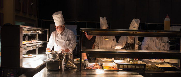 Group of chefs in white uniforms and hats working in a professional kitchen at Wrest Point Hotel & Conference Centre. They are preparing and plating food under bright lights on a stainless steel counter, with various kitchen utensils and equipment visible. The dark background contrasts with the well-lit workspace.