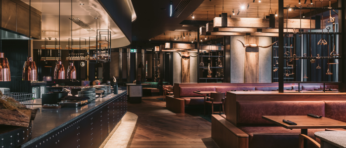 Interior of an upscale restaurant at Wrest Point Hotel & Conference Centre featuring brown leather booths, wooden tables, and a warm-toned design. The ceiling has wooden beams with hanging lights, and an open kitchen area with copper pots and pans is visible on the left. Decorative longhorn skulls adorn the walls, contributing to the sophisticated ambiance.