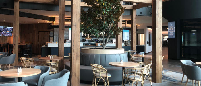 Interior of a modern café or restaurant at Wrest Point Hotel & Conference Centre featuring wooden beams, a central potted tree with cushioned seating, round tables with wicker and cushioned chairs, and a bar area with stools.