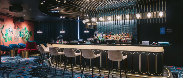 Stylish bar at Wrest Point Hotel & Conference Centre featuring a curved counter with gold accents, high chairs, and a display of bottles behind the bar. The ceiling has an artistic light arrangement, the floor is covered with patterned carpet, and colorful abstract wall art is visible on the left.