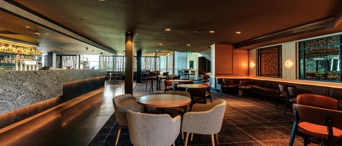 Modern bar and lounge area at Wrest Point Hotel & Conference Centre featuring round tables with cushioned chairs, booth seating, and a sleek stone bar counter. The space has dim lighting, contemporary decor, and large windows allowing natural light to enhance the warm ambiance.