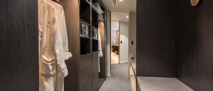 Modern walk-in closet at Wrest Point Hotel & Conference Centre featuring dark wood paneling, white bathrobes hanging on the left, and shelves with compartments including safes. The carpeted floor leads to an open doorway revealing a connected bedroom or living area.
