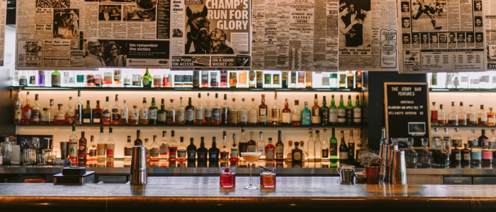 MACq01 Eat and Drink 1 2025 Well-stocked bar with liquor bottles on shelves, two drinks on the counter, a cocktail shaker, and bar tools. Newspaper clippings and headlines are displayed above the bar, creating a vintage ambiance.