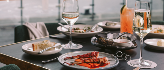MACq01 Eat and Drink 2 2025 Elegant dining table with plates of grilled octopus skewers in red sauce, oysters or shellfish, and bread slices. Drinks include two glasses of white wine and an orange cocktail with a lemon slice. The background features large windows with a scenic, out-of-focus view.